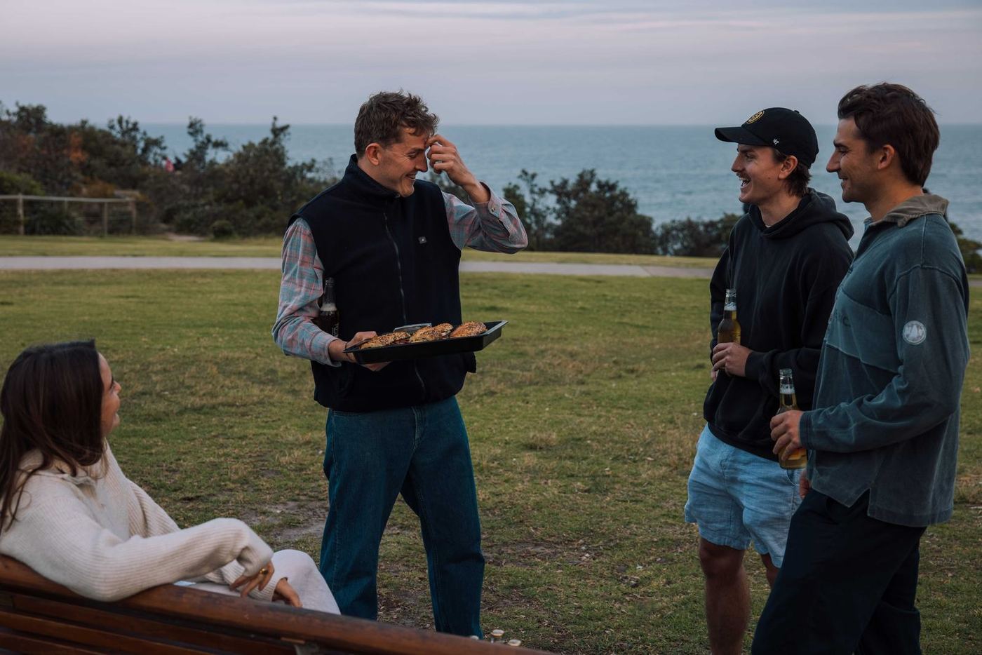 Friends laughing over a tray of pesto sandwiches on a clifftop above the ocean