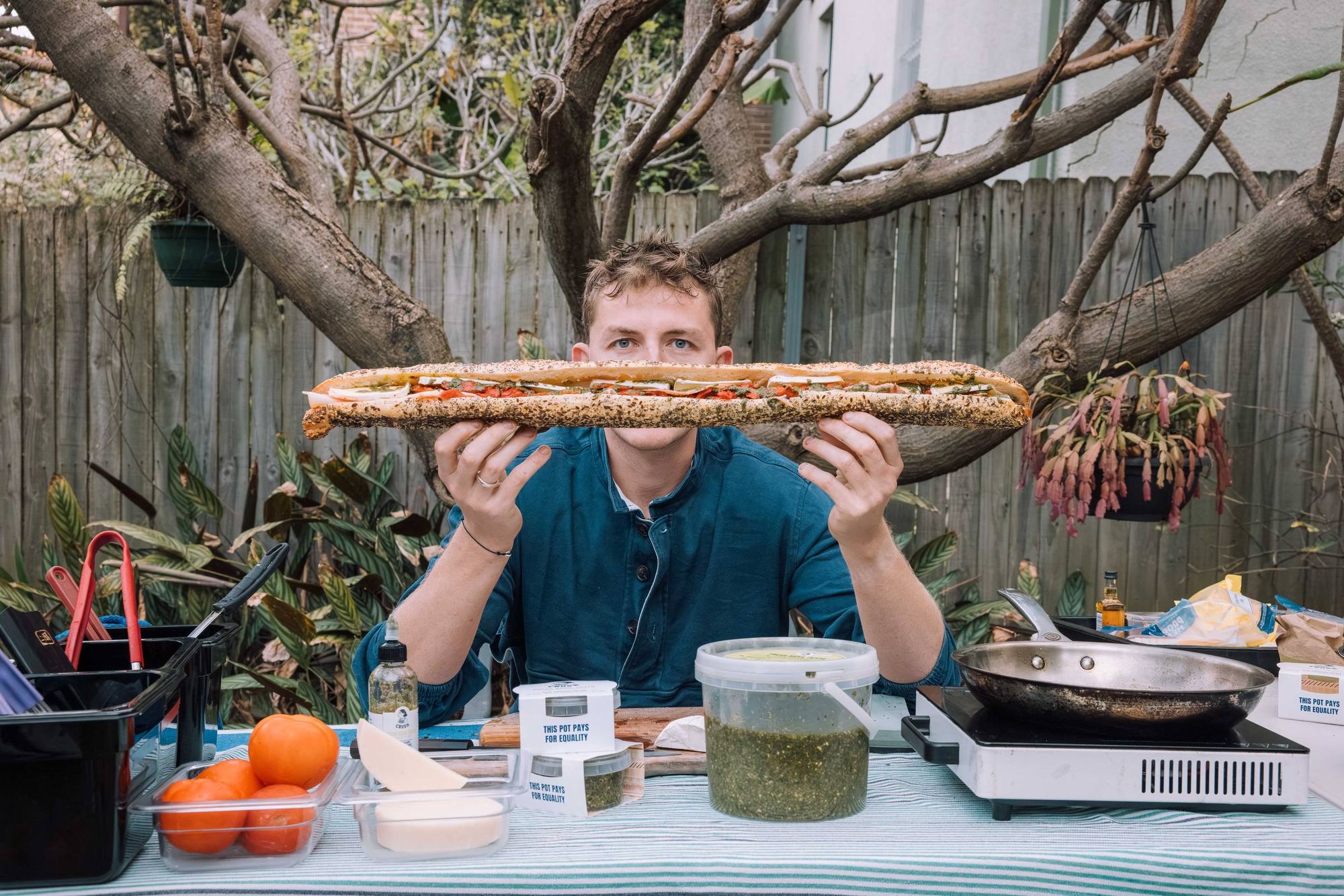 Ed, Crush's founder, holding a long pesto baguette across his face behind an outdoor cooking table