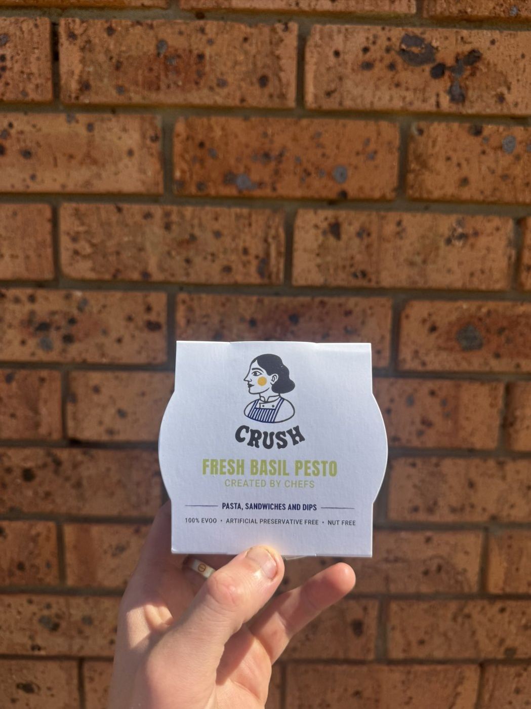 A hand holding the Crush Fresh Basil Pesto 150ml retail pot against a brick wall