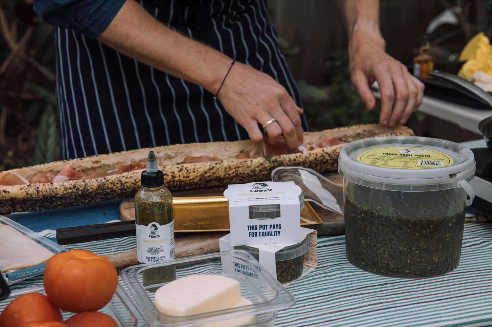 A chef building a long prosciutto baguette beside Crush pots, a squeeze bottle and a 2kg tub