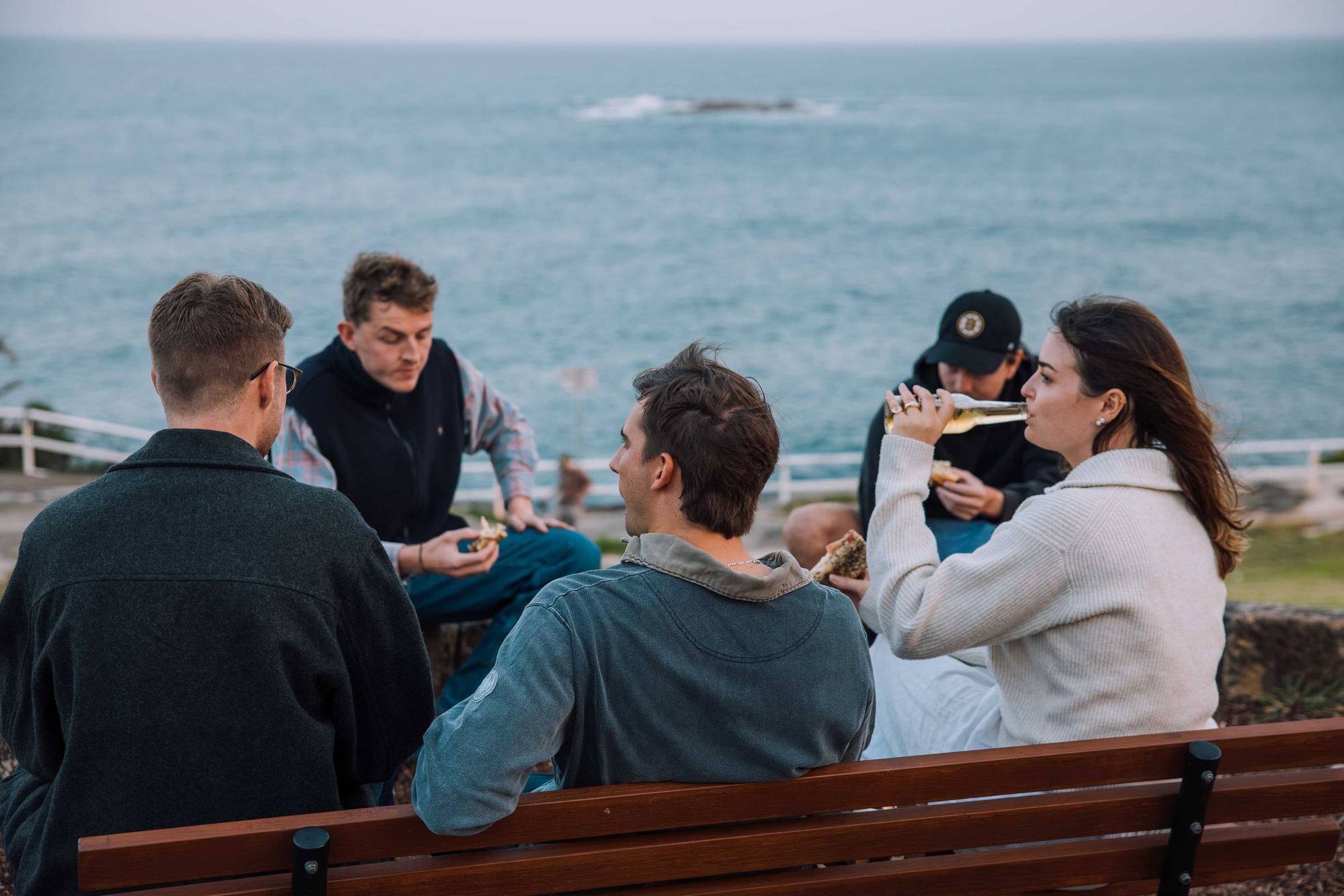 Friends sharing food on a clifftop bench above the ocean
