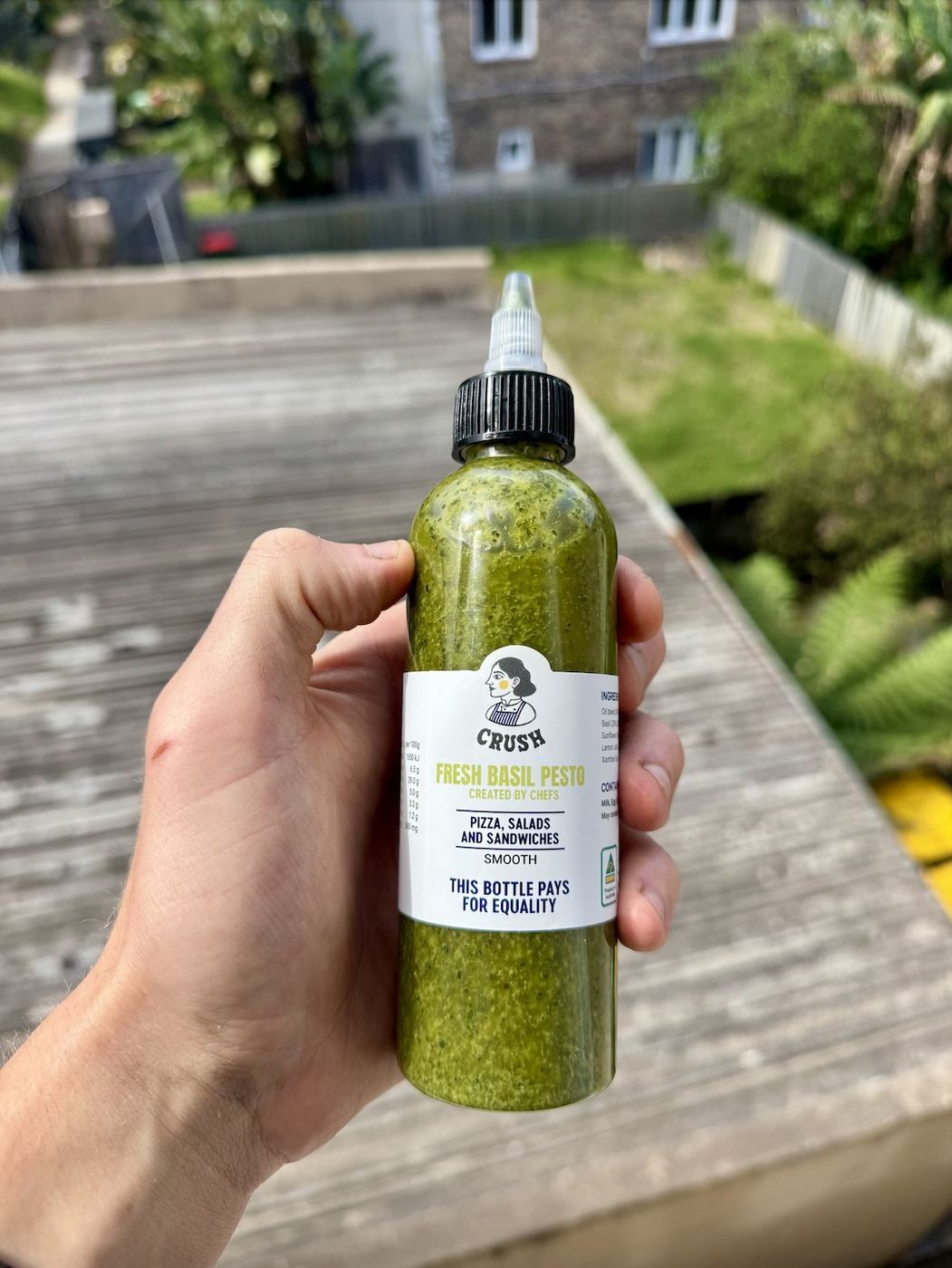 A hand holding a Crush Fresh Basil Pesto squeeze bottle labelled This bottle pays for equality